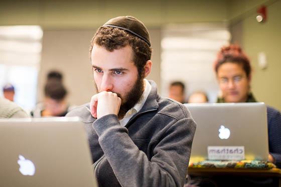Photo of a male student in a yarmulke, 坐在波胆网站的演讲厅里，看着他的电脑